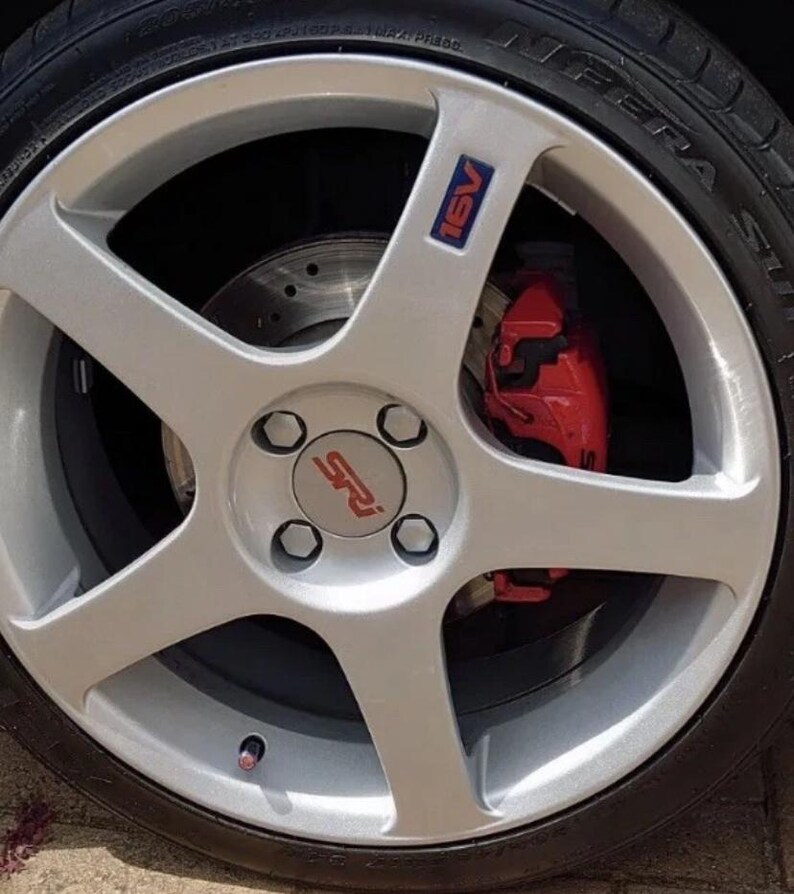 May include: A silver alloy wheel with a five-spoke design. The wheel features a red brake caliper and a tire. The center cap has the letters "SRI" in red. The wheel also has a blue and red "16V" emblem.