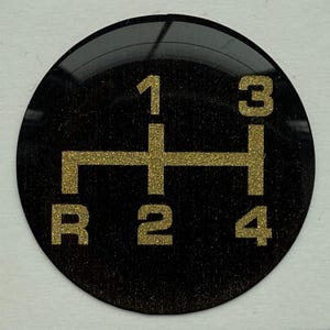Puede incluir: Un patrón de cambio de marchas negro y circular con números y letras dorados con purpurina. El patrón incluye los números 1, 2, 3 y 4, y la letra R para la marcha atrás. El diseño es un patrón en H clásico.
