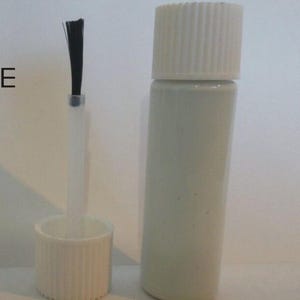 Könnte beinhalten: Eine weiße Flasche mit geripptem Verschluss und Pinselapplikator. Der Pinsel hat schwarze Borsten. Die Flasche ist zylindrisch und enthält wahrscheinlich eine Flüssigkeit, möglicherweise Nagellack oder ein ähnliches Kosmetikprodukt.
