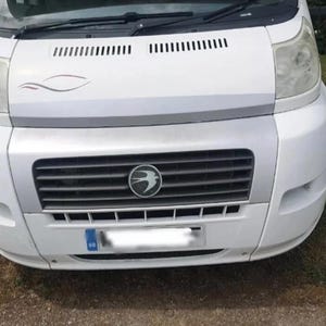 Puede incluir: Autocaravana blanca con sección frontal plateada. La parte delantera presenta una parrilla con un emblema circular, faros y una matrícula. El vehículo está estacionado sobre hierba.