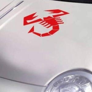 May include: A bright red scorpion decal applied to the white hood of a vehicle. The graphic features a stylized, geometric representation of a scorpion with its tail curved upward, positioned centrally on the smooth, curved surface of the car hood.