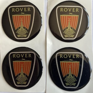 Könnte beinhalten: Vier schwarze, runde Rover-Emblem-Aufkleber. Jeder Aufkleber zeigt das Wort "ROVER" in Weiß oben, über einem Schilddesign mit einem roten und goldenen Wikingerschiff. Die Aufkleber befinden sich auf einer weißen Rückseite.