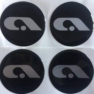 Może przedstawiać: Cztery czarne, okrągłe naklejki z błyszczącym wykończeniem. Każda naklejka przedstawia szarą, stylizowaną literę "A" z małym kółkiem. Naklejki są ułożone w siatce 2x2 na białym tle.