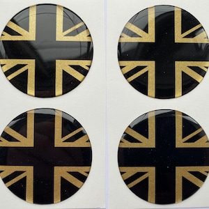 Puede incluir: Cuatro pegatinas redondas y brillantes de color negro con un diseño de la bandera Union Jack en dorado. Las pegatinas están dispuestas en una cuadrícula de 2x2 sobre un fondo blanco. El diseño dorado de la bandera destaca sobre el negro.