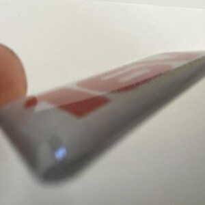 May include: A close-up of a rectangular, transparent sticker with red text on a grey background. The sticker is held between fingers, with the text partially visible. The sticker has a slight curve.