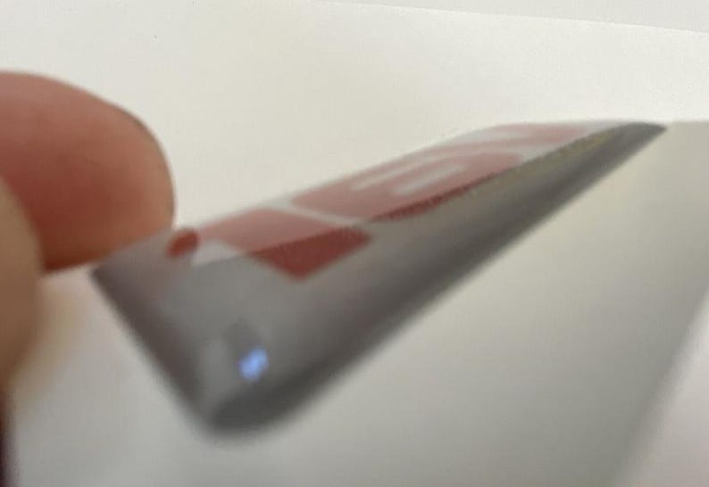 May include: A close-up of a rectangular, transparent sticker with red text on a grey background. The sticker is held between fingers, with the text partially visible. The sticker has a slight curve.