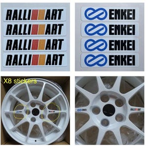 May include: A collection of automotive stickers featuring the "Ralli Art" and "Enkei" logos. The "Ralli Art" stickers have a black background with the logo in white and a red, orange, and yellow stripe. The "Enkei" stickers have a white background with a blue logo and black text. Also shown are white car rims with the stickers applied.