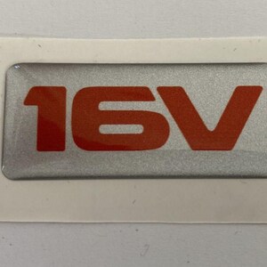 May include: A rectangular silver sticker with the text "16V" in bright orange. The sticker has a silver background and is surrounded by a white border. The sticker is likely for a vehicle.