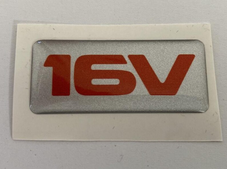 May include: A rectangular silver sticker with the text "16V" in bright orange. The sticker has a silver background and is surrounded by a white border. The sticker is likely for a vehicle.