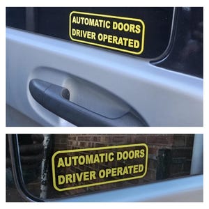 May include: Two yellow rectangular stickers with black text that read "AUTOMATIC DOORS DRIVER OPERATED" are affixed to the windows of a vehicle. The stickers are on a silver vehicle door, near the door handle.