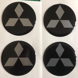 May include: Four black circular decals with a gray three-diamond logo. The decals are glossy and appear to be stickers or emblems. The logo is a stylized geometric design.