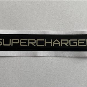 Puede incluir: Un emblema rectangular negro con la palabra "SUPERCHARGED" en letras plateadas. El emblema está sobre un respaldo blanco, lo que sugiere que es una pegatina o calcomanía para un vehículo. El diseño es simple y moderno.
