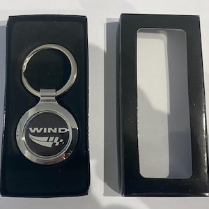 May include: A silver keyring with a black circular emblem displaying the word "WIND" and a stylized design. The keyring is presented in a black gift box with a clear window.