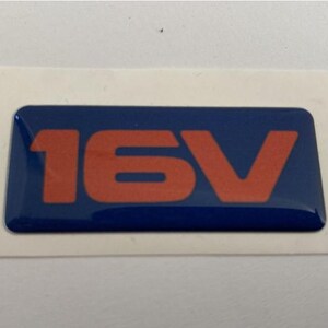 May include: A blue rectangular sticker featuring the orange text '16V'. The sticker has a glossy finish and is set against a white background. The text is in a bold, sans-serif font.