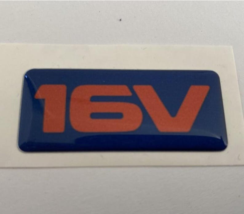 May include: A blue rectangular sticker featuring the orange text '16V'. The sticker has a glossy finish and is set against a white background. The text is in a bold, sans-serif font.