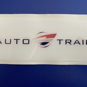 May include: A rectangular white sticker with the words "AUTO TRAIL" in black, with a stylized logo featuring red, silver, and black elements. The sticker is on a white backing, with a blue background.
