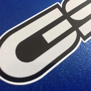 May include: A sticker with a white and black design. The sticker has a rounded shape with a black interior and white outline. The background is a solid blue colour.