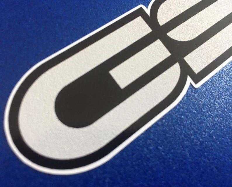 May include: A sticker with a white and black design. The sticker has a rounded shape with a black interior and white outline. The background is a solid blue colour.