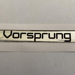 Może przedstawiać: Srebrna prostokątna odznaka z czarnym napisem "Vorsprung" bezszeryfowym. Odznaka znajduje się na białym tle.