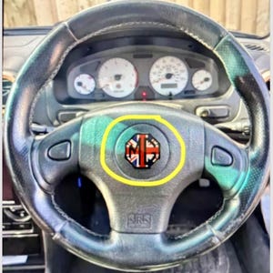 May include: A black leather steering wheel with a silver center console and dashboard. The center of the steering wheel features a badge with the letters "MG" and a Union Jack design. The dashboard has white gauges.