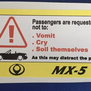 May include: A yellow sticker with a red warning triangle and a black car silhouette. Text reads: "Passengers are requested not to: Vomit, Cry, Soil themselves. As this may distract the pilot." The text "MX-5" is also present.