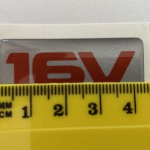 May include: A rectangular sticker with the text "16V" in red against a silver background. The sticker is on a white background. A yellow ruler is placed below the sticker, showing measurements in centimetres.