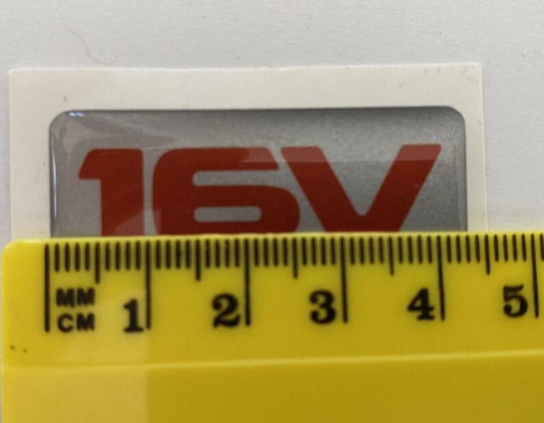 May include: A rectangular sticker with the text "16V" in red against a silver background. The sticker is on a white background. A yellow ruler is placed below the sticker, showing measurements in centimetres.