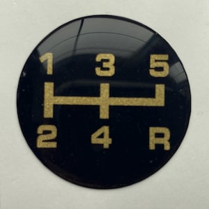 Może przedstawiać: Czarny, okrągły schemat zmiany biegów ze złotymi cyframi i schematem zmiany biegów. Widoczne są cyfry 1, 2, 3, 4, 5 i R, wskazujące pozycje biegów.
