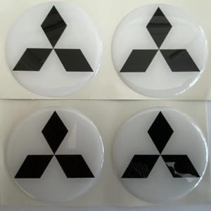 May include: Four white, circular stickers featuring the Mitsubishi logo. The logo consists of three black diamond shapes arranged in a triangular pattern. The stickers are on a white background.
