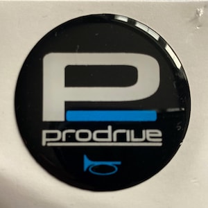 Pode incluir: Um autocolante redondo preto com a palavra "prodrive" em texto branco. Um grande "P" branco está acima do texto, com uma linha horizontal azul. Um gráfico de buzina azul na parte inferior.