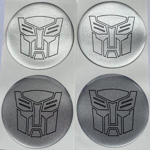 Op de afbeelding: Vier ronde zilverkleurige stickers gerangschikt in een twee-bij-twee raster. Elke sticker bevat een zwarte lijntekening van het Autobot-insigne uit de Transformers-franchise, gekenmerkt door een gestileerd ontwerp van een robotgezichtsmasker.