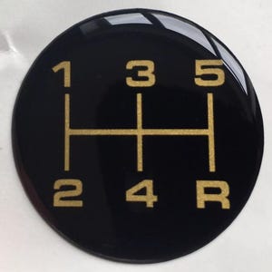 May include: A black, circular gear shift knob with gold-colored numbers and lines indicating the gear positions: 1, 2, 3, 4, 5, and R (reverse). The knob has a glossy finish.