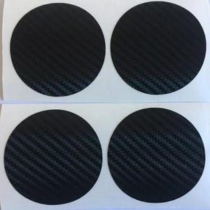 Könnte beinhalten: Vier schwarze, kreisförmige Aufkleber mit einem Carbonfaser-Webmuster. Die Aufkleber sind in zwei Reihen zu je zwei Stück auf einem weißen Träger angeordnet. Das Carbonfaser-Design hat eine strukturierte Optik.