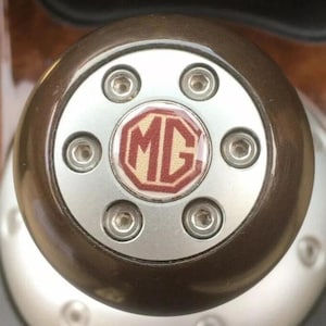 Pode incluir: Close-up de um botão de mudança de marcha castanho e prateado. O botão tem um design circular com um centro prateado e um anel exterior castanho. O centro apresenta o logótipo MG vermelho e bege, fixado por seis parafusos.