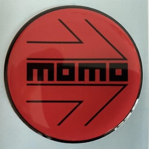 Pode incluir: Um autocolante redondo vermelho com uma borda preta. O autocolante apresenta a palavra "MOMO" em letras pretas, com desenhos de setas pretas apontando para fora. Provavelmente para uso automotivo, o autocolante tem um design arrojado.