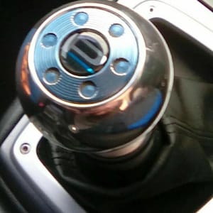 May include: Shiny chrome gear shift knob with a blue accent ring and blue detailing. The knob is round with a black and blue center. The shift boot is black.