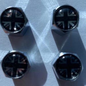 Puede incluir: Cuatro tuercas de rueda cromadas con un diseño de la Union Jack. Cada tuerca tiene una cabeza hexagonal con el diseño de la bandera en blanco y negro en el centro. Las tuercas están dispuestas en forma cuadrada, proyectando sombras sobre una superficie clara.