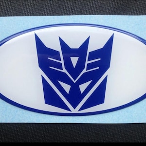 May include: An oval-shaped sticker featuring the Decepticon logo. The emblem is a stylized, blue, geometric design set against a white background, framed by a blue border. The sticker is likely for automotive or personal use.