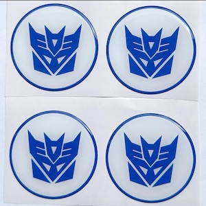 Peut inclure: Quatre autocollants circulaires disposés en une grille de deux par deux sur une feuille de support blanche. Chaque autocollant présente un logo Decepticon bleu centré sur un fond circulaire blanc, entouré d'une fine bordure circulaire bleue.