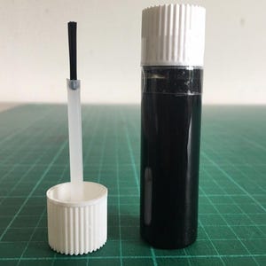 May include: A small, clear bottle filled with black liquid, next to a white cap and applicator brush. The bottle has a white cap. The applicator brush has a black bristle tip. The items are on a green grid surface.