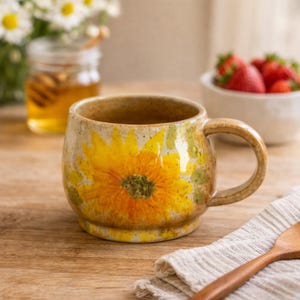 Peut inclure: Une tasse en céramique avec un grand motif de tournesol jaune. La tasse a une forme arrondie, une anse robuste et une finition tachetée et terreuse. En arrière-plan, un pot de miel, des fleurs et des fraises.