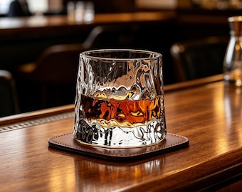 May include: A clear, textured whiskey glass filled with amber liquid, resting on a square brown leather coaster. The glass sits on a polished wooden bar, with a blurred background of a bar setting.