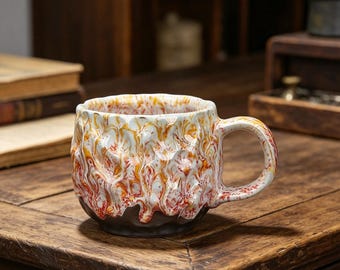 May include: A ceramic mug with a textured, lava-like design in white, orange, and red hues. The mug has a dark brown base and a curved handle. It sits on a wooden surface, showcasing its unique, handcrafted appearance.