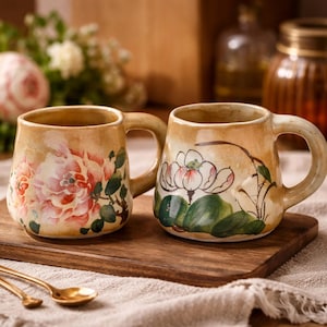 Könnte beinhalten: Zwei Keramikbecher mit floralen Designs. Ein Becher zeigt eine große rosa Pfingstrose, der andere eine weiße Lotusblume mit grünen Blättern. Beide Becher haben einen beigen Hintergrund und gebogene Griffe. Sie stehen auf einem Holzbrett.