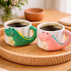 Puede incluir: Dos tazas de cerámica con diseños de caimanes. Una taza es verde con detalles amarillos, la otra es rosa con detalles azules. Ambas tazas tienen un fondo blanco con rayas grises verticales y están llenas de café oscuro.