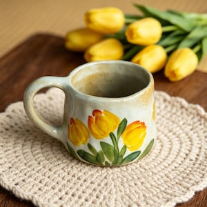 May include: A ceramic mug with a pale blue glaze, decorated with hand-painted yellow tulips with orange accents and green leaves. The mug rests on a cream crocheted coaster, with yellow tulips in the background.