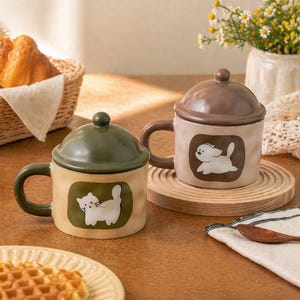 Könnte beinhalten: Zwei Keramikbecher mit Deckel, einer grün und einer braun, jeweils mit einem Cartoon-Tierdesign. Der grüne Becher hat eine weiße Katze, und der braune Becher hat einen weißen Hund. Beide Becher haben passende Griffe und stehen auf einer Holzoberfläche.