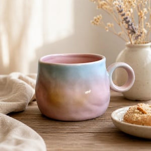 May include: A ceramic mug with a pastel colour gradient of pink, blue, and yellow. The mug has a rounded shape with a large handle. A small plate with a biscuit and a vase with dried flowers are in the background.