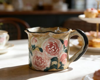 May include: A ceramic mug with a beige background and a black handle. The mug is decorated with pink roses and green leaves. The rim of the mug has a wavy design.