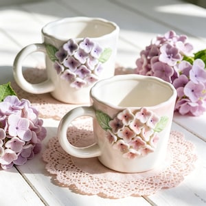 May include: Two white ceramic mugs with a floral hydrangea design in shades of pink and purple. The mugs have a rounded handle and sit on pink lace doilies. The background includes fresh hydrangea blooms.